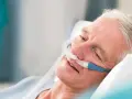 Älterer Mann mit Nasenkanüle, der entspannt im Bett liegt. Die Sauerstoffzufuhr mit **high flow** hilft ihm beim Atmen.