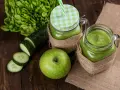 Dwa słoiki z zielonym smoothie, obok jabłko, plasterki ogórka i sałata. Orzeźwiający drink z ogórkiem na drewnianym tle.