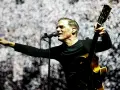 Bryan Adams w Polsce, śpiewa i gra na gitarze na tle świetlistej scenografii.