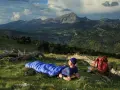 Mann liegt in blauer Biwakschachtel im Freien, umgeben von Berglandschaft. Rucksack und Wanderstöcke daneben.