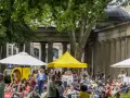 Menschen genießen entspannte **Veranstaltungen am Wochenende** im Freien vor einer Säulenfassade. Sonnenschirme und Zelte spenden Schatten.