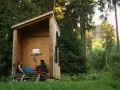 Zwei Personen entspannen sich in einer einfachen Holzhütte, bereit für wild camping. Umgeben von hohen Bäumen und üppigem Grün.