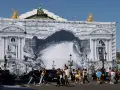 Des passants traversent devant l'Opéra Garnier, dont la façade est recouverte d'une bâche monumentale simulant des travaux et une arche rocheuse.