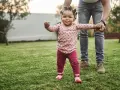 Ein kleines Mädchen mit lustigen Zöpfen macht seine ersten Schritte im Gras, gehalten von einer erwachsenen Hand. Wann laufen Babys? Heute ist ein guter Tag dafür!