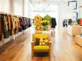 Interior de un showroom de ropa con percheros llenos de prendas coloridas y un sillón verde con cojines estampados.