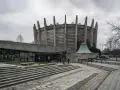 Monumentalny budynek w stylu brutalizm z betonowymi żebrami i szklaną fasadą. Schody prowadzą do wejścia, a wokół zielone trawniki.
