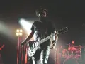 Un guitariste charismatique d'un groupe de rock mythique, en pleine performance sur scène, éclairé par des projecteurs.
