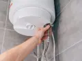 Mano che controlla la valvola scaldabagno perde. Tubi flessibili collegati a un boiler bianco su sfondo piastrellato.