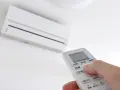 Mano ajustando el **timer on aire acondicionado** con el control remoto. El aire acondicionado blanco está montado en la pared.