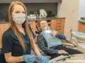 Uśmiechnięta pacjentka daje kciuk w górę podczas pierwszej wizyty u dentysty po latach. Dentystka w masce i rękawiczkach przygotowuje narzędzia.