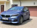 Niebieskie BMW 218i zaparkowane przed domem z brązową bramą garażową.