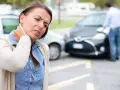 Kobieta z bólem pleców po wypadku samochodowym trzyma się za szyję. W tle widać uszkodzone samochody.