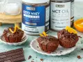 Pyszne keto muffiny z mąki migdałowej, polane czekoladą i ozdobione skórką pomarańczową. Obok stoją puszki z odżywką białkową i olejem MCT.