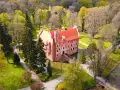 Zamek na wodzie w Wojnowicach otoczony zielenią i drzewami. Czerwona bryła zamku z charakterystycznym dachem wznosi się nad fosą.
