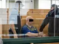 Skruszony gangster w kajdankach, prowadzony przez policjantów w sali sądowej.