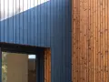 Drewniana elewacja z ciemnogranatową wiatroizolacją i naturalnym drewnem. Okno z ciemną ramą.