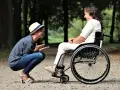 Opiekun osoby niepełnosprawnej pochyla się, by porozmawiać z kobietą na wózku inwalidzkim w parku.