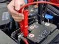 Hand klemmt rote Überbrückungsklemme an die Autobatterie, um Plus und Minus zu verbinden.