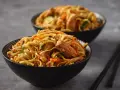 Zwei Schüsseln mit gebratenen Nudeln, Karotten und Fleisch. Ein Paar Essstäbchen liegt daneben.
