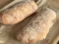 Dwa świeżo upieczone bochenki ciabatty, posypane mąką, leżą na papierze do pieczenia. Idealne do wypróbowania tego ciabatta przepis!