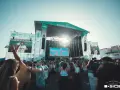 Multitud en concierto, con pantallas gigantes mostrando a artistas. El sol brilla sobre el escenario "Estrella de Levante" del festival B-Side.