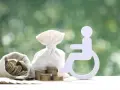Symbol osoby na wózku inwalidzkim obok worków z pieniędzmi i monet. Świadczenie wspierające wyrównanie szans.