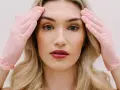 Kobieta z blond włosami w różowych rękawiczkach dotyka skroni. Przygotowanie do zabiegu, jakim jest liposukcja twarzy, by uzyskać idealny kontur.