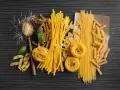 Różnorodne rodzaje makaronu, od cienkich nitek po tagliatelle i fusilli, ułożone na ciemnym tle. Wartości odżywcze makaronu zależą od jego składu.