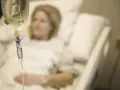 Infusion vorbereiten: Eine Frau liegt im Krankenhausbett, während eine Infusionstropfflasche und ein Tropfschlauch im Vordergrund zu sehen sind.