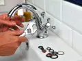 Main tenant utilise une clé pour changer un joint de robinet. Des joints sont posés sur le lavabo.