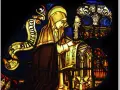 Stained glass depicts a cloaked figure, likely the head of a monastery, diligently writing on a scroll.