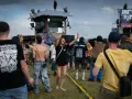 Une foule danse sous la pluie à un festival, une femme au centre lève les bras. On aperçoit une scène et des bannières, peut-être un dj français anime l'événement.