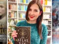 Uśmiechnięta kobieta trzyma książkę "The Atlas Six". W tle widać półki z książkami, w tym inne serie książek fantasy.