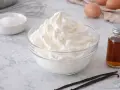 Un bol de chantilly maison aérienne, avec des ingrédients pour la préparer : œufs, vanille, sucre glace et un fouet.