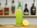 Zielony, orzeźwiający drink Tokyo Iced Tea z plasterkiem cytryny i metalową słomką, na tle półki z butelkami alkoholu.