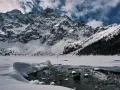 Zamarznięte Morskie Oko, otoczone ośnieżonymi szczytami Tatr. W oddali widać ludzi spacerujących po lodzie, a na pierwszym planie płynie strumyk.