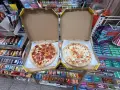 Dwie pizze w pudełkach, jedna z salami, druga serowa. Obok półki ze słodyczami i napojami w sklepie Żabka.