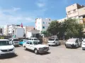 Taxis warten auf Fahrgäste in einer sonnigen Stadt in Marokko. Weiße Gebäude und Bäume säumen die Straße.