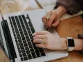 Dłonie piszące na klawiaturze laptopa, z widocznym zegarkiem i pierścionkami.