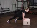 Kobieta wykonuje ćwiczenie mountain climbers na skrzyni. W tle siłownia z ciężarami.