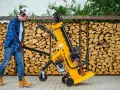 Mężczyzna w kasku i okularach ochronnych przygotowuje się do pracy z szybką łuparką do drewna. Za nim piętrzą się stosy drewna opałowego.