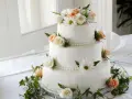 Elegancki tort weselny ozdobiony białymi i brzoskwiniowymi różami, margaretkami i zielonymi liśćmi. Piękne, jakie żywe kwiaty na tort!