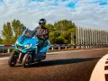 Niebieski Peugeot Metropolis 400 z kierowcą w kasku pędzi po zakręcie drogi, otoczony zielenią i błękitnym niebem.