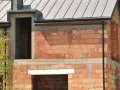 Budynek w budowie z cegły, widoczne okna i drzwi, dach pokryty blachą. Widać cofnięte nadproże do rolet.