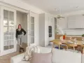 Kobieta w eleganckim stroju stoi w przestronnym salonie z aneksem kuchennym. Wnętrze w stylu hygge zachwyca jasnymi kolorami, drewnianymi elementami i przytulnymi dodatkami.