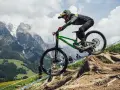 Troy Brosnan in mid-air on a green downhill bike, with a dramatic mountain backdrop.