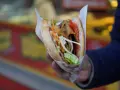 Kebab w bułce z warzywami i mięsem, trzymany w dłoni.