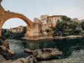 Iconic Stari Most bridge over the Neretva River, a must-see for any Bosnia travel adventure. People watch from the bridge as swimmers enjoy the emerald waters below.