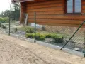 Montaż siatki ogrodzeniowej przy drewnianym domu. Widać zielone słupki, siatkę i betonowy podmurówkę.