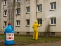 Person en tenue jaune effectuant un nettoyage facade eau de javel sur un mur sale. Une grande bouteille d'eau de javel est posée au sol.
